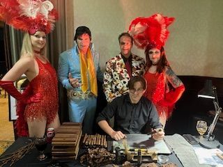 Costumed performers pose around a seated man at a table with papers and props indoors.