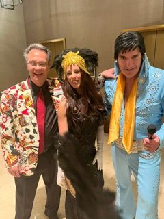 Three costumed people posing indoors, one holding a microphone, with floral, feathered, and blue outfits.