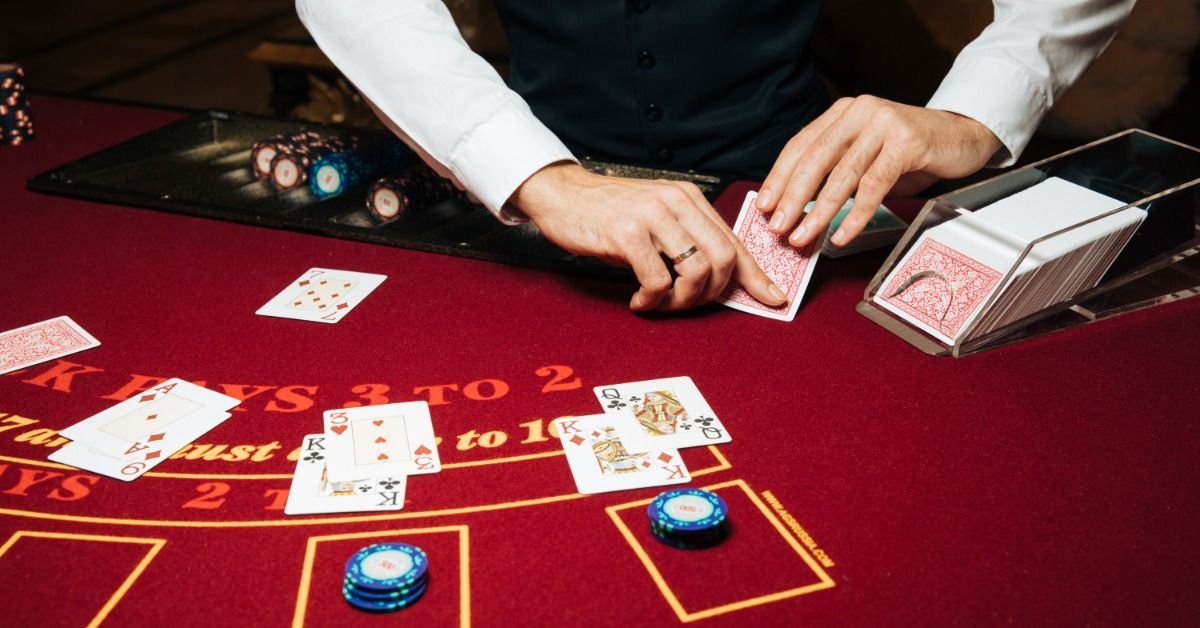 A croupier in a white shirt and dark vest deals playing cards on a red blackjack table.