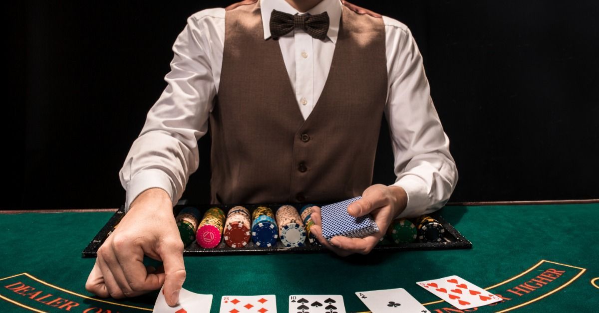 A Hold'em dealer in a bow tie and vest hands out playing cards on a green casino table. He has rows of chips.