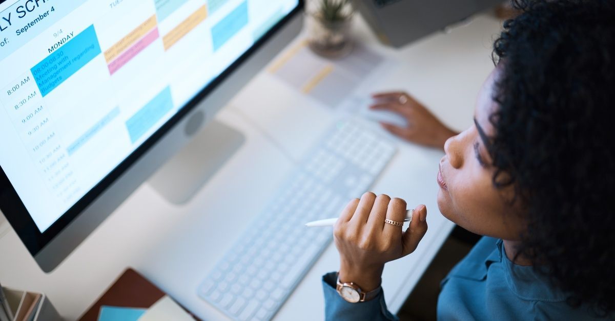 A woman holding a pen sits at a desk with a computer. She looks at a monthly planning calendar on her screen.