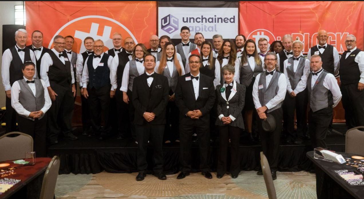 Group of people in formal attire posing on a stage at an Unchained event with orange backdrop and banner