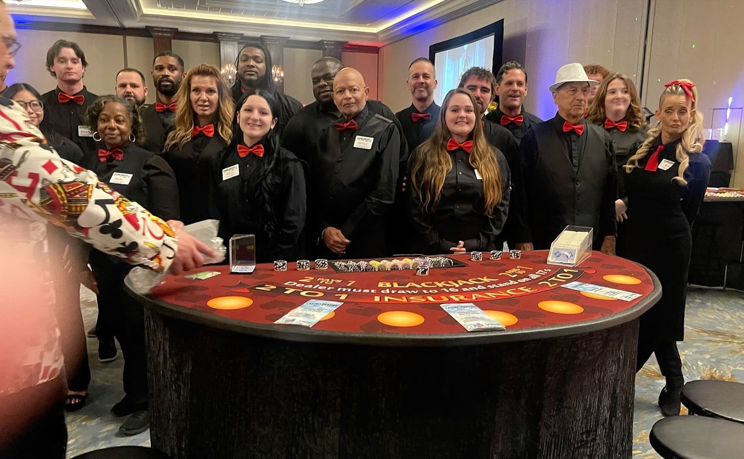 A group of people wearing black uniforms and red bowties stand behind a semicircular casino table in a conference room.