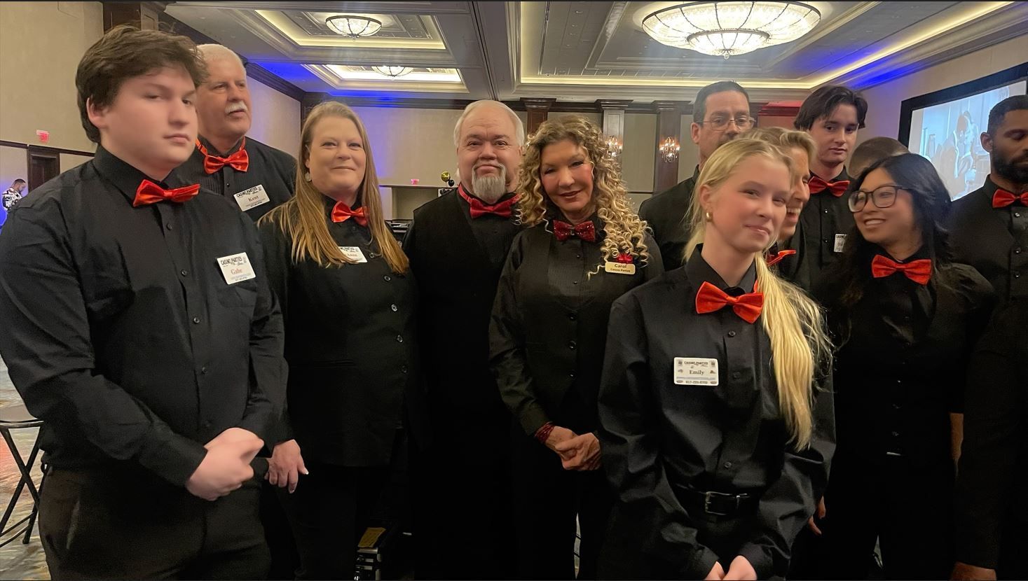 A group in black shirts and red bow ties poses for a photo in a large, brightly lit banquet hall.
