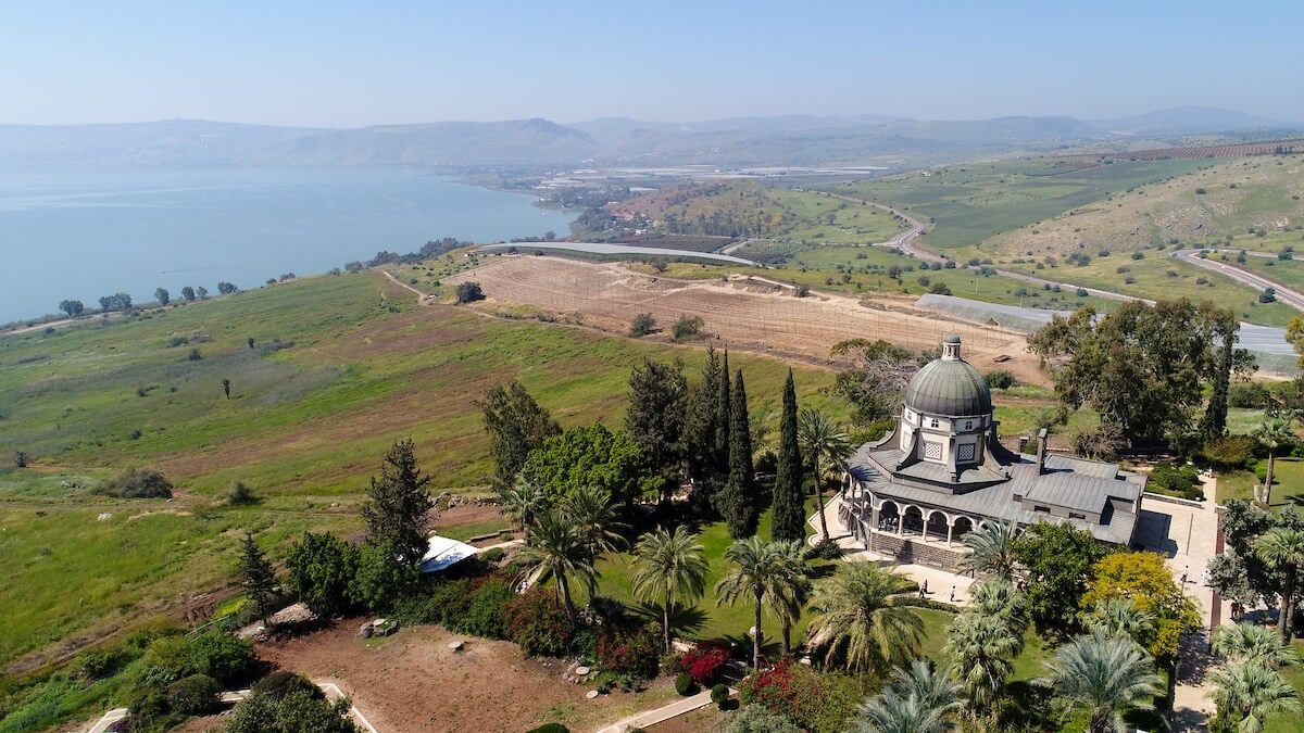 Coastal hillside with a domed shrine, trees, farmland, and distant sea under a hazy sky