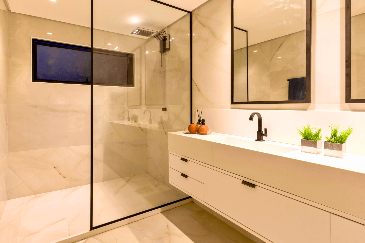 Baño moderno con ducha con mampara de cristal, tocador flotante blanco y espejos con marco negro.