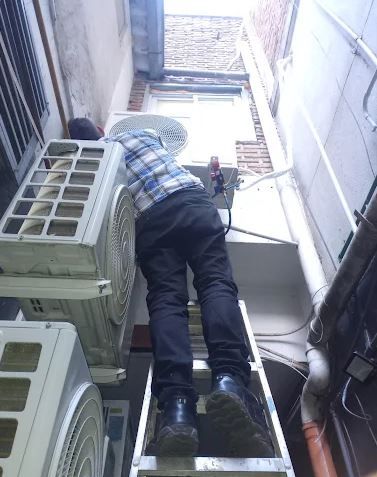 Una persona sube por una escalera metálica en un callejón estrecho junto a unidades de aire acondicionado y tuberías apiladas.