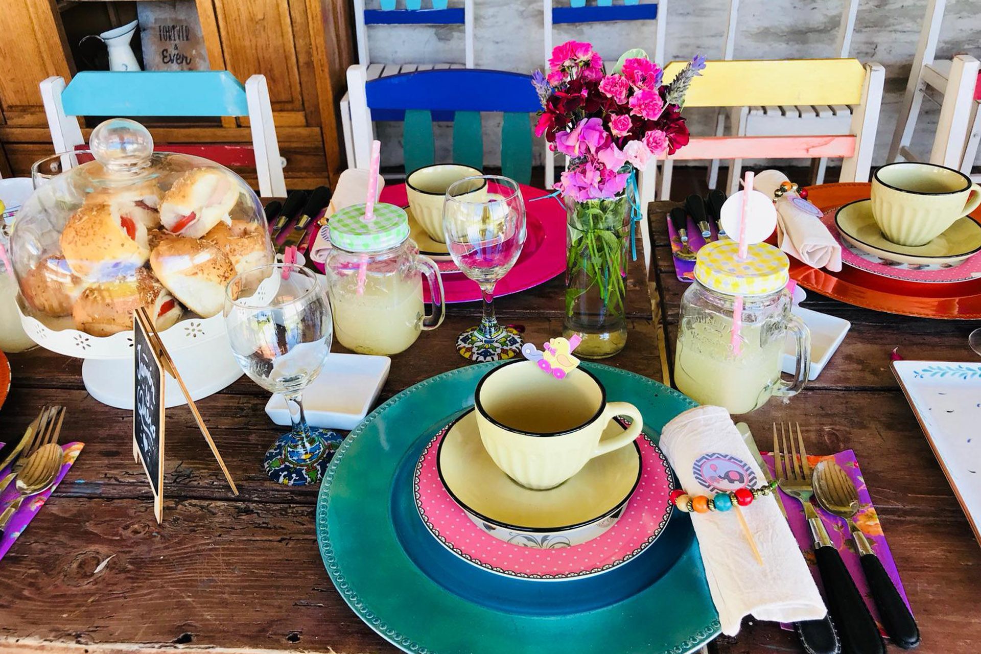 Una mesa rústica preparada para una merienda con comida dispuesta en varios niveles, tazas, platos coloridos, bebidas en tarros de cristal y arreglos florales.