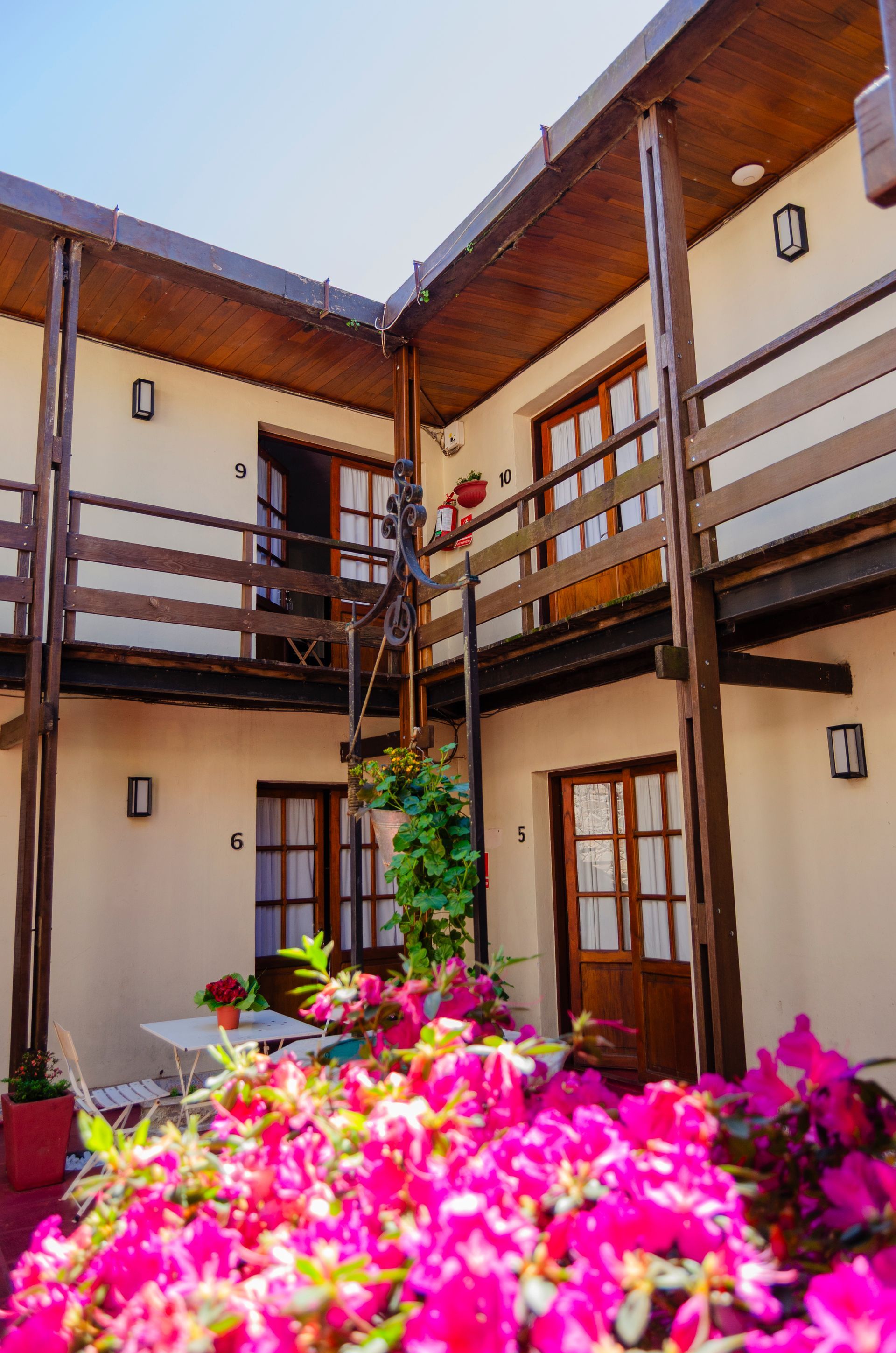 Patio de un edificio de dos plantas con balcones de madera, paredes de colores claros, puertas de cristal y flores de color rosa intenso en la fachada.