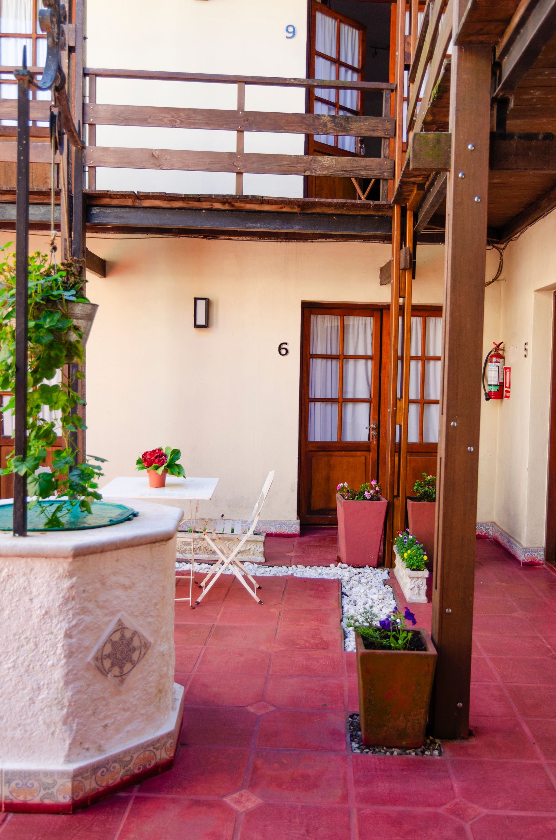 Un patio con suelo de baldosas rojas, un pozo de piedra, plantas en macetas y una pequeña mesa de bistró frente a las habitaciones marcadas con los números 5, 6 y 9.