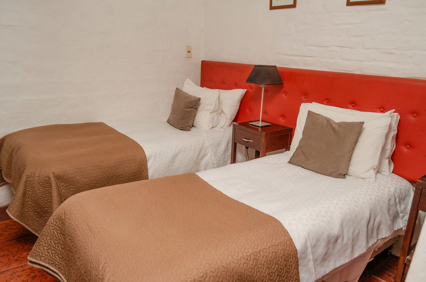 Dos camas individuales con mantas marrones, sábanas blancas y almohadas grises, frente a un cabecero acolchado de color rojo brillante, en una habitación blanca.