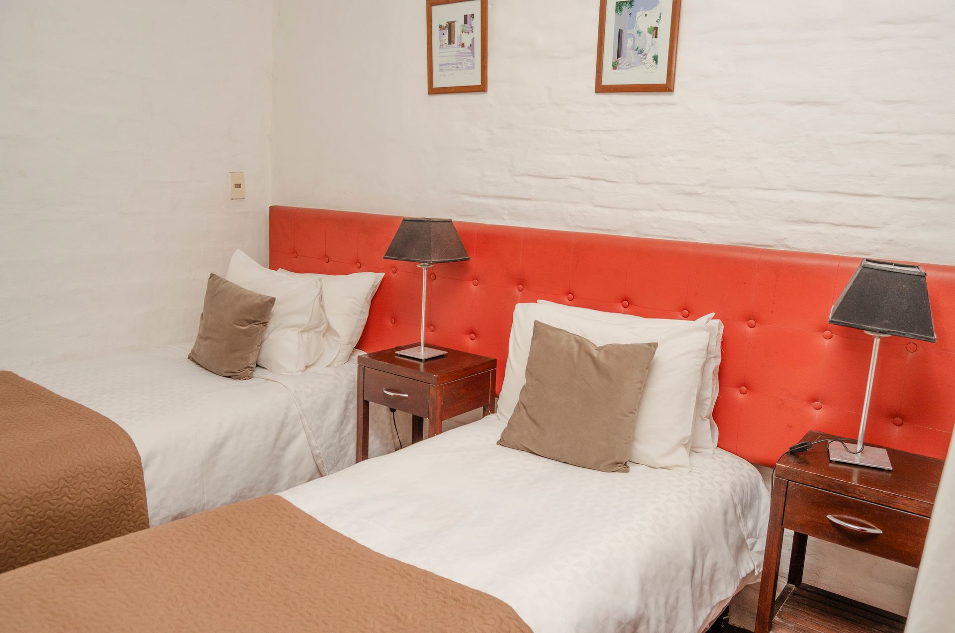 Dos camas individuales con sábanas blancas, almohadas marrones y mesitas de noche de madera oscura, con un cabecero acolchado de color naranja, en una habitación de hotel.