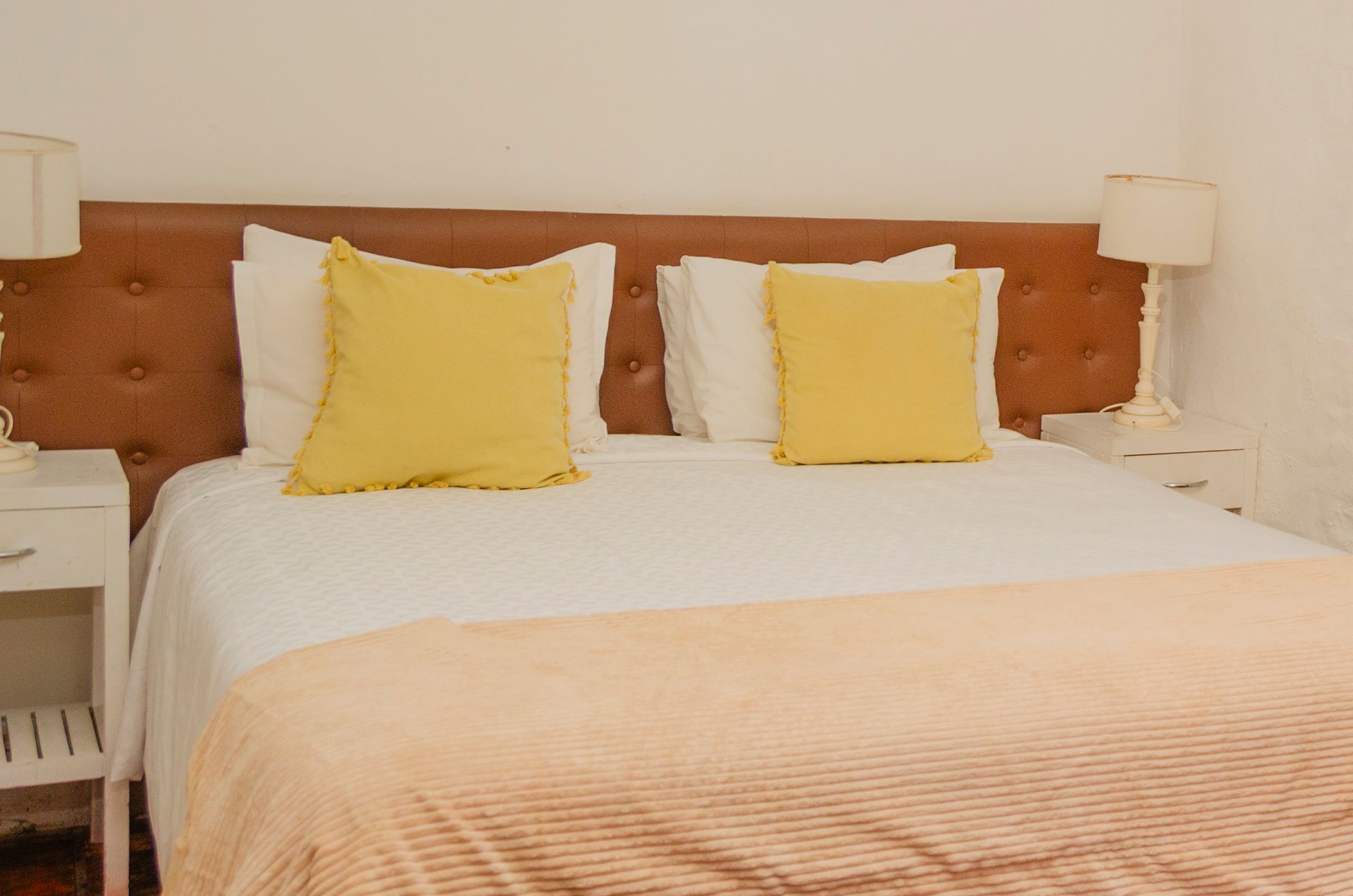 Una cama con cabecero acolchado de cuero marrón, sábanas blancas, cojines decorativos amarillos y una manta texturizada de color melocotón claro.