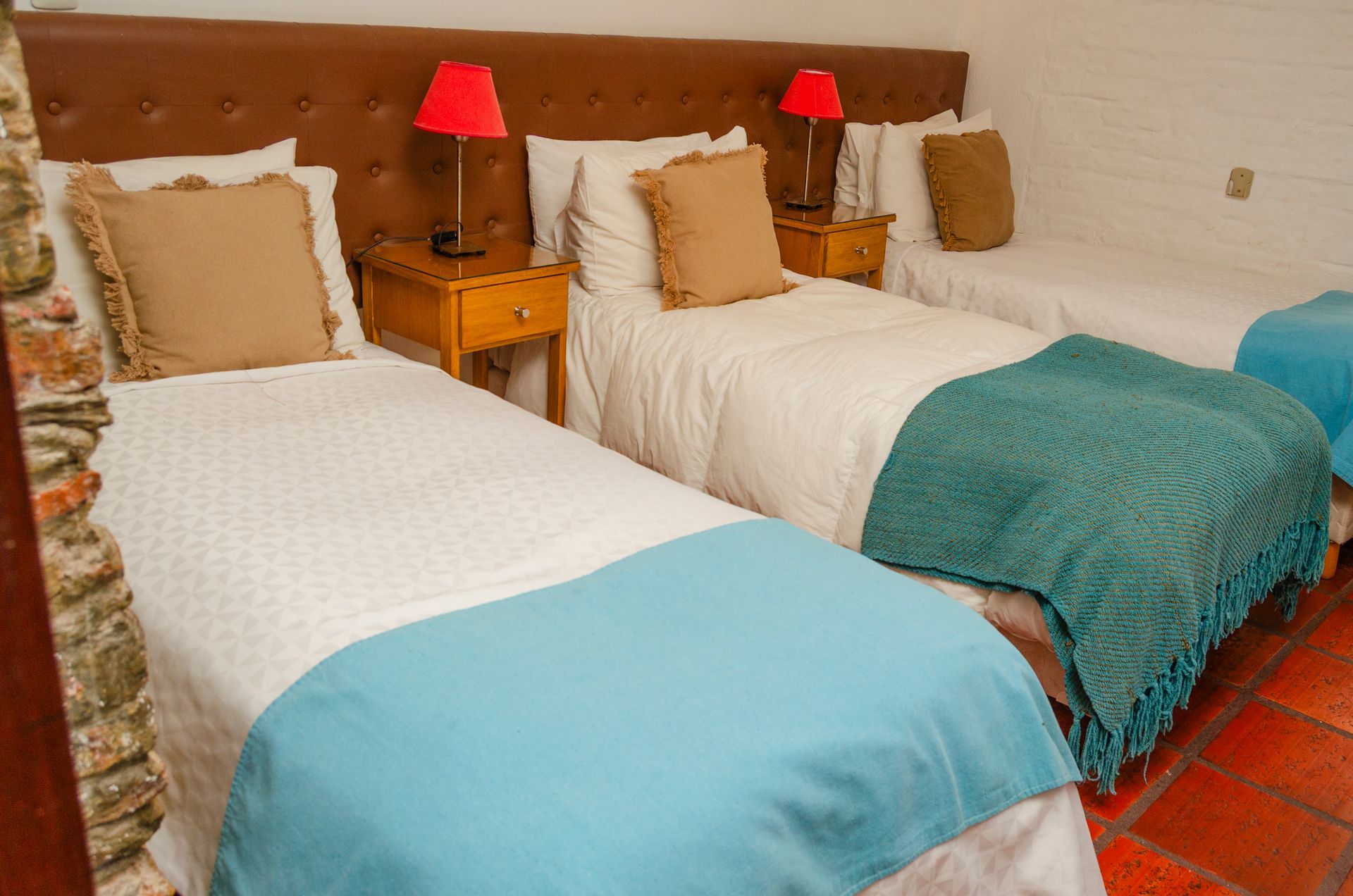 Tres camas en una habitación con cabecero tapizado de color marrón, mesitas de noche de madera, lámparas rojas y mantas decorativas de color azul claro y verde azulado.