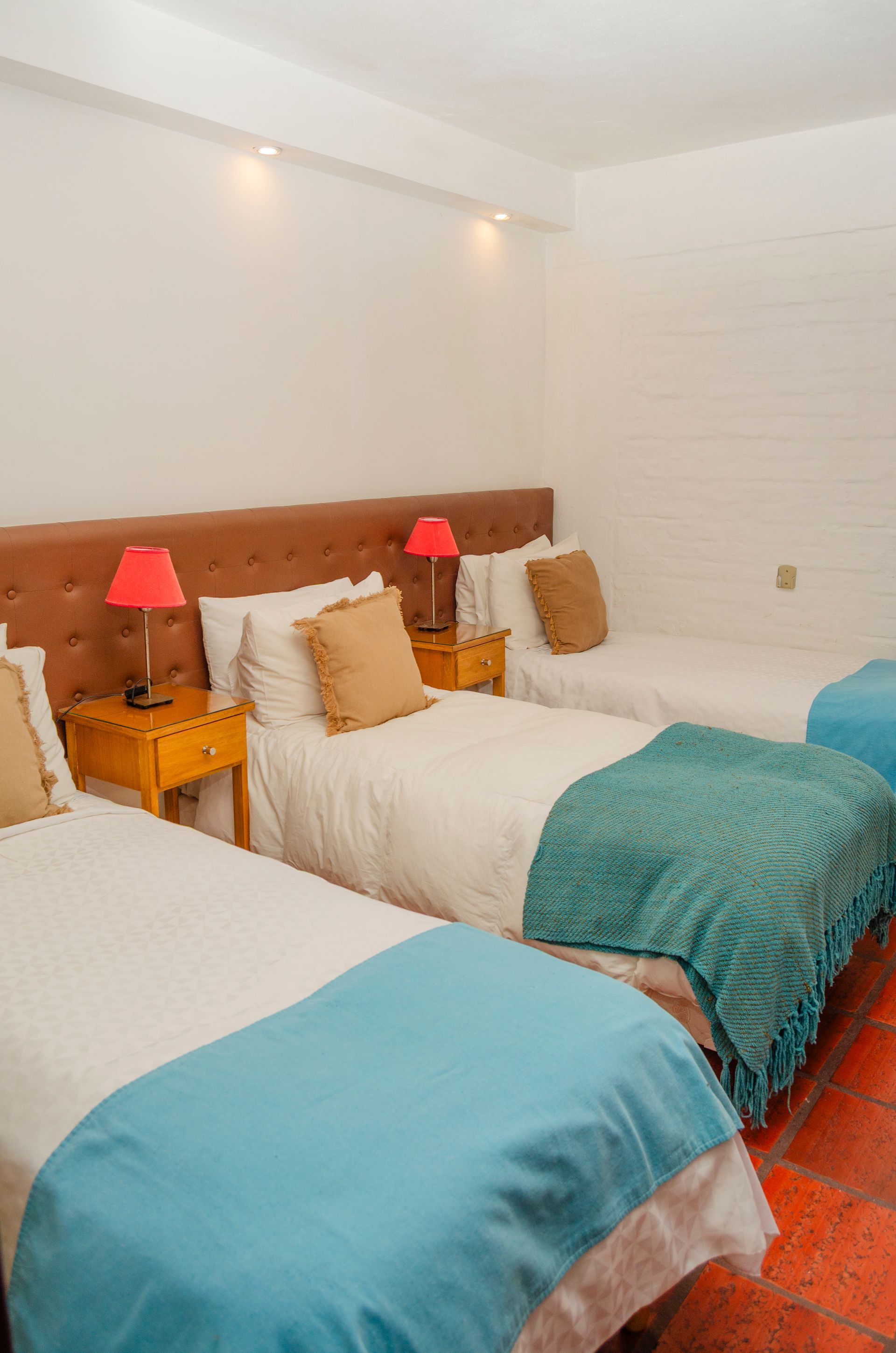 Un dormitorio con tres camas individuales con sábanas blancas, mantas turquesas, almohadas color canela y mesitas de noche amarillas.
