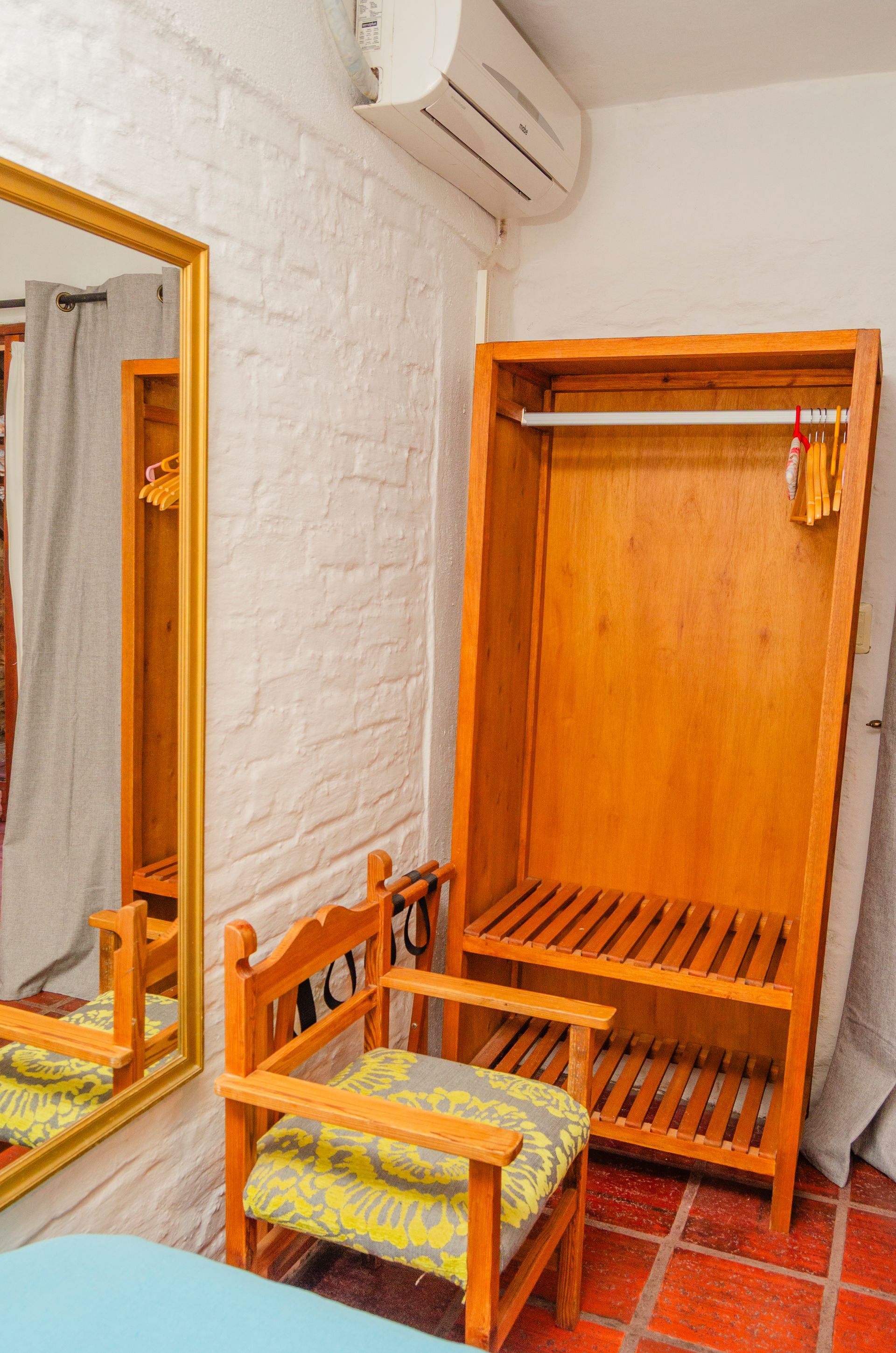 Un armario de madera y una silla a juego con tapicería estampada se encuentran junto a un espejo de cuerpo entero contra una pared de ladrillo.