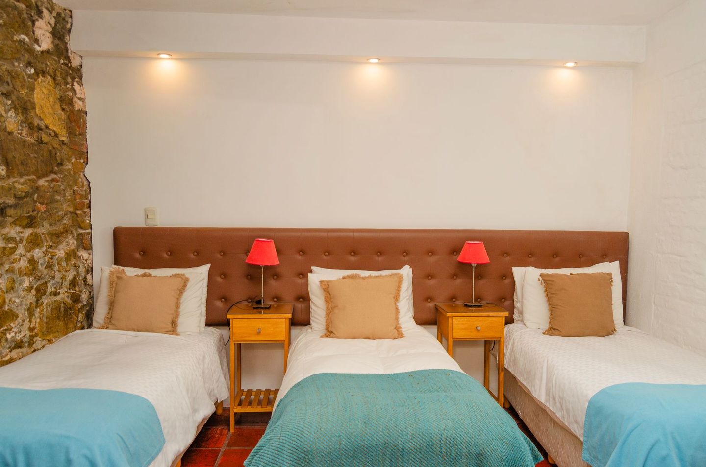 Tres camas individuales con sábanas blancas y mantas color turquesa, dispuestas frente a un cabecero marrón, en una habitación con pared de piedra.