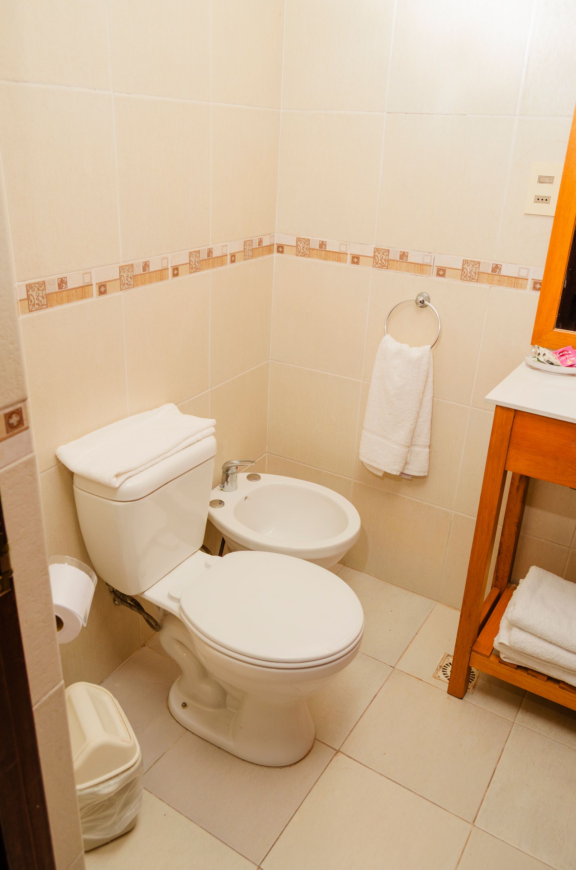 Un baño limpio con azulejos de color beige, inodoro blanco, bidé, toallero con una toalla blanca y lavabo de madera.