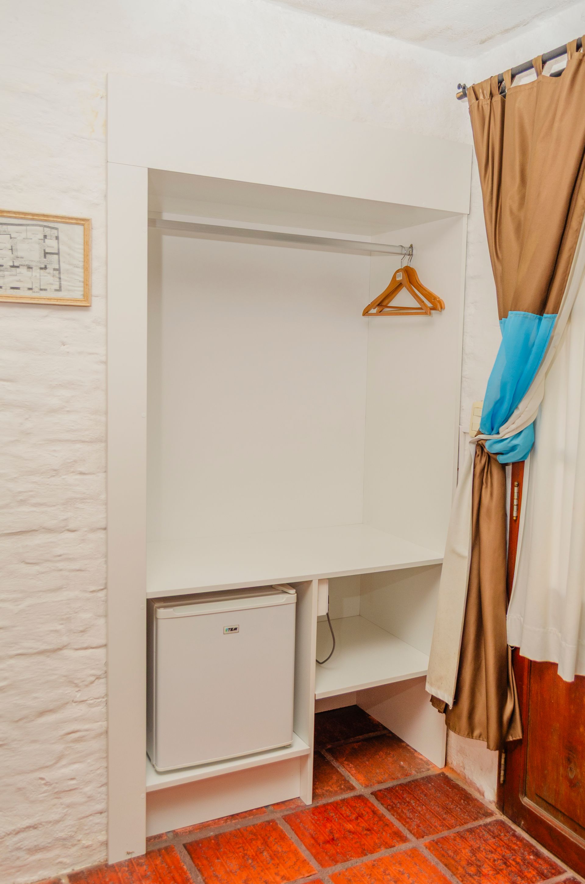 Un armario empotrado blanco con barra para colgar ropa, un pequeño frigorífico blanco y una estantería, situados en una habitación con suelo de baldosas.