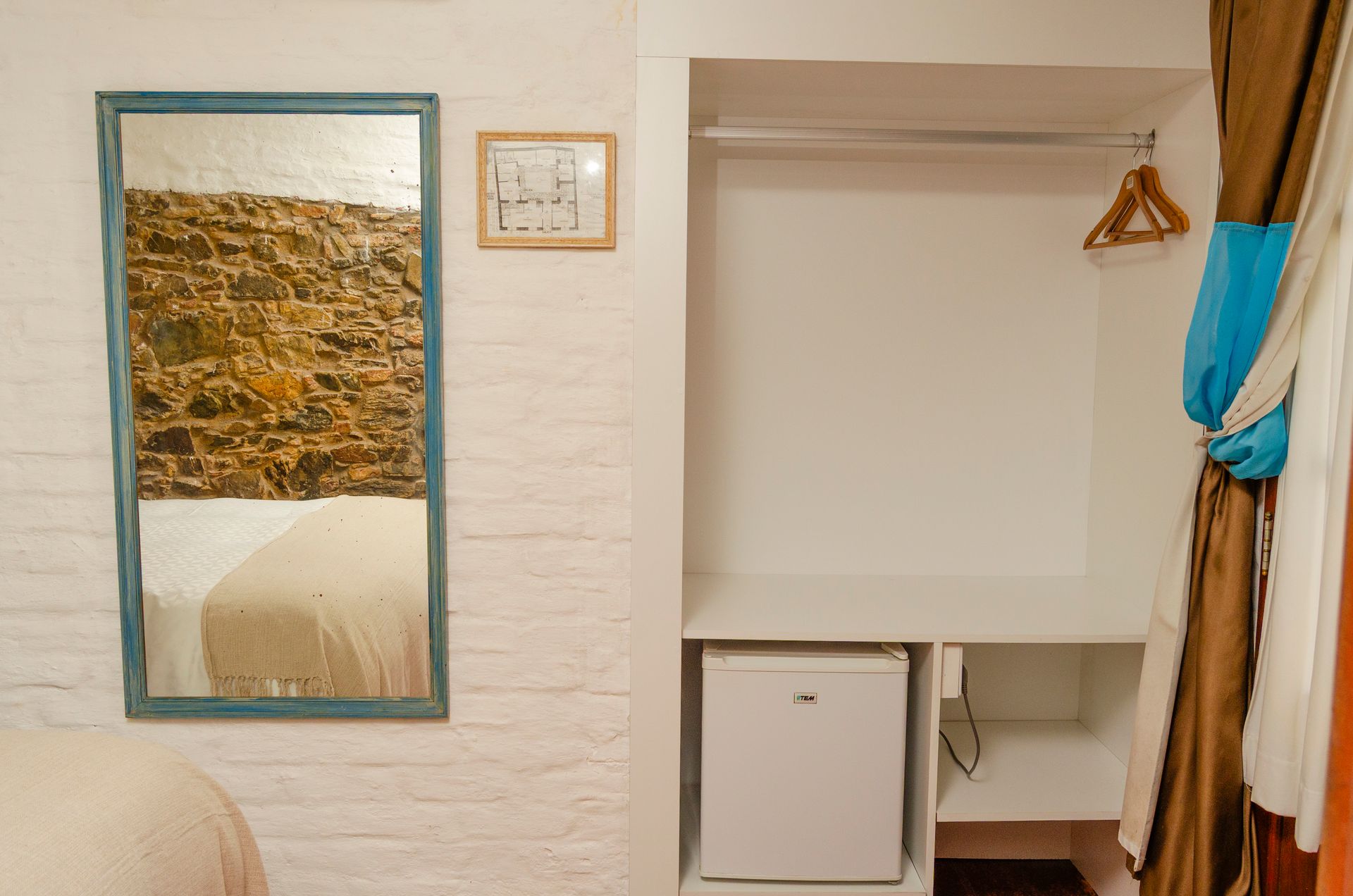 Una pared blanca presenta un espejo enmarcado que refleja una pared de piedra, una pequeña nota enmarcada y un armario empotrado con un minibar.