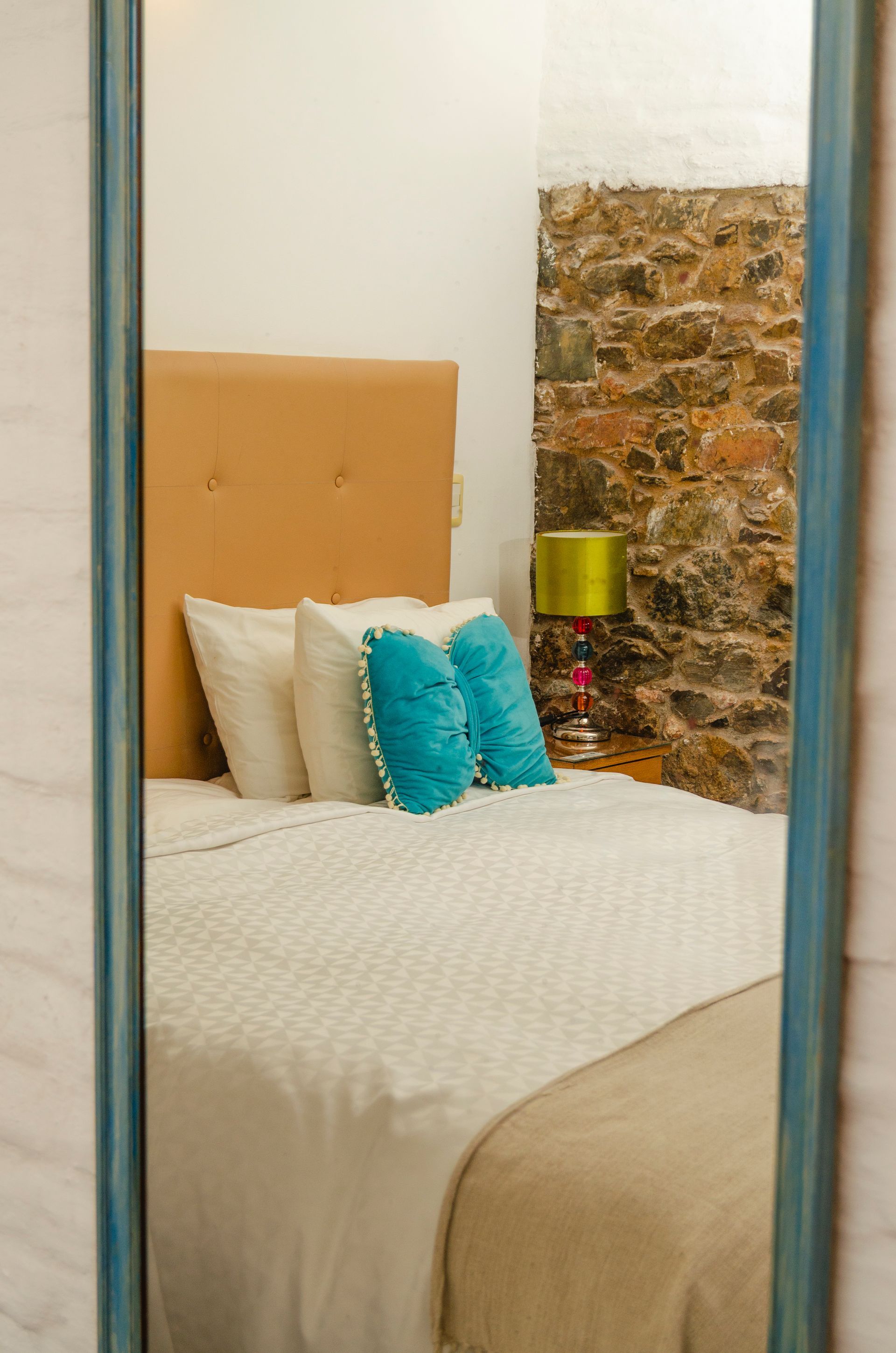 Vista a través de un marco de espejo de madera que muestra una cama con ropa de cama blanca, almohadas color turquesa y una pared de piedra en una habitación rústica.