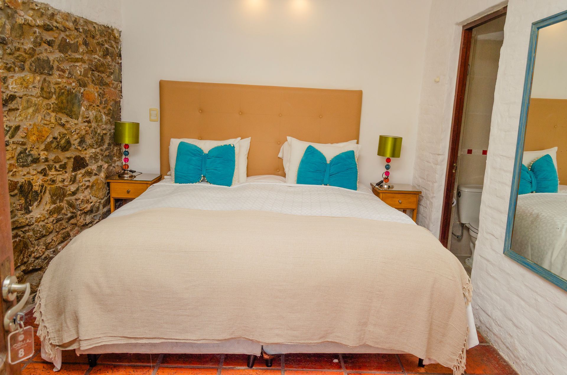 Un dormitorio con una cama king size, una pared de piedra texturizada, un cabecero beige, almohadas azules y un gran espejo que refleja la habitación.