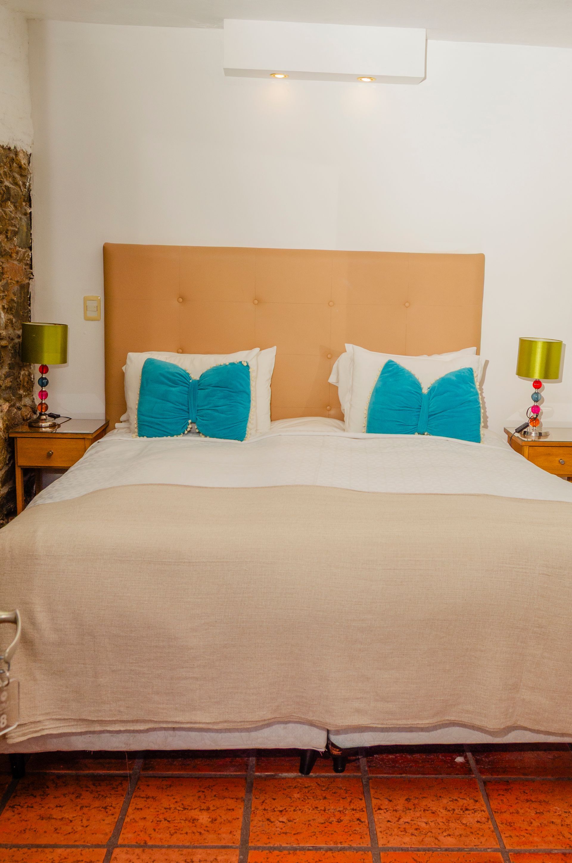 Una cama con cabecero color tostado, ropa de cama beige y dos almohadas de color verde azulado brillante en forma de pajarita, flanqueada por lámparas verdes a juego.
