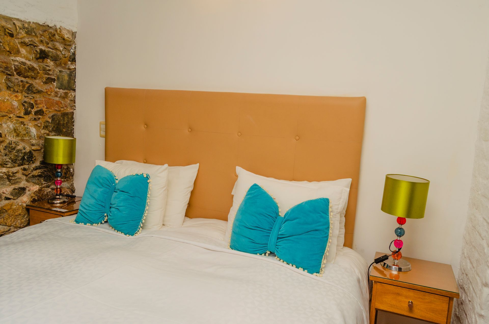 Una cama con cabecero color canela, ropa de cama blanca y dos almohadas azul brillante en forma de lazo, flanqueada por lámparas sobre mesitas de noche de madera.