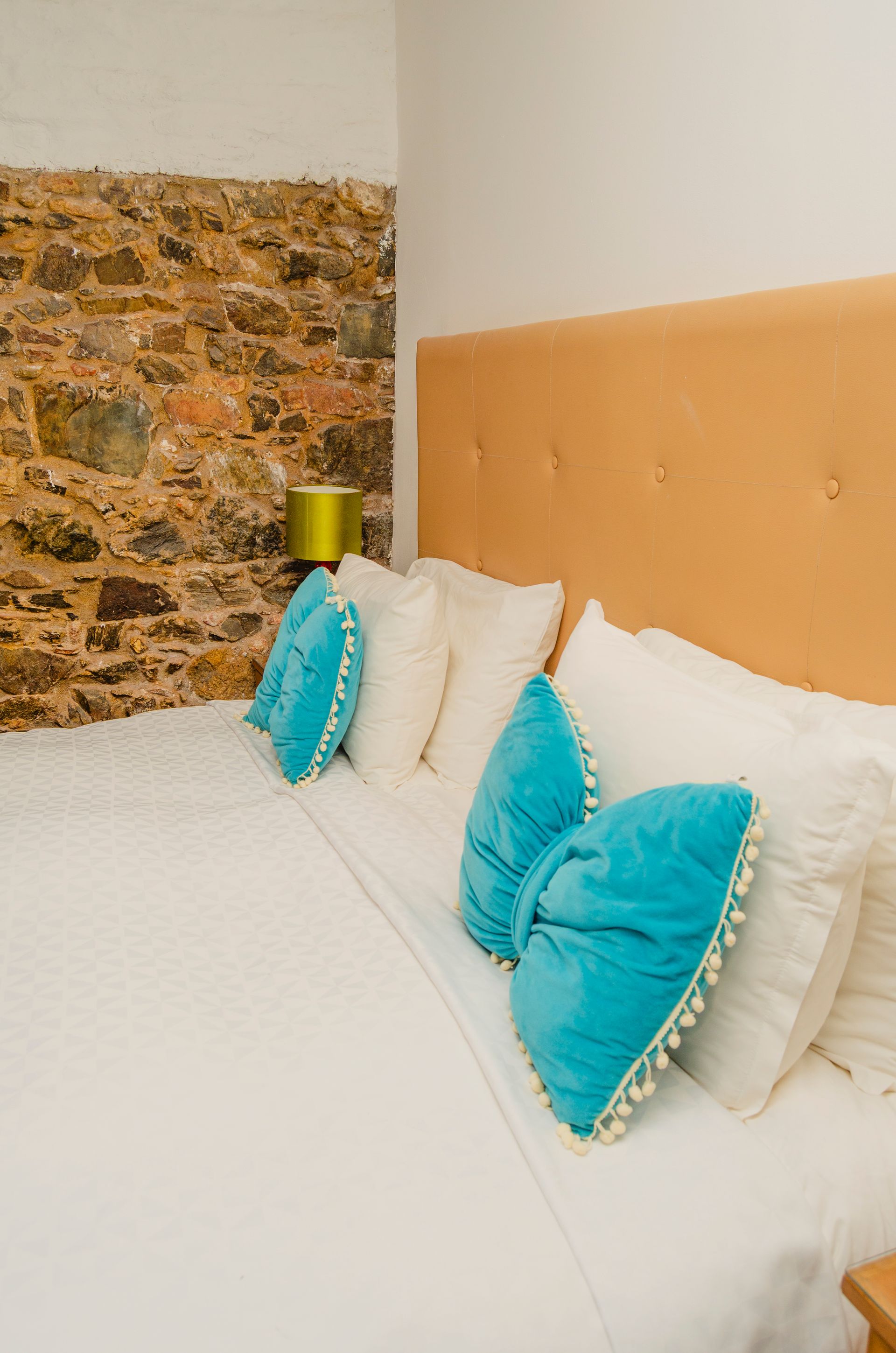 Una cama hecha con cabecero de color beige, sábanas blancas y cojines decorativos de color verde azulado contra una pared de piedra rústica.