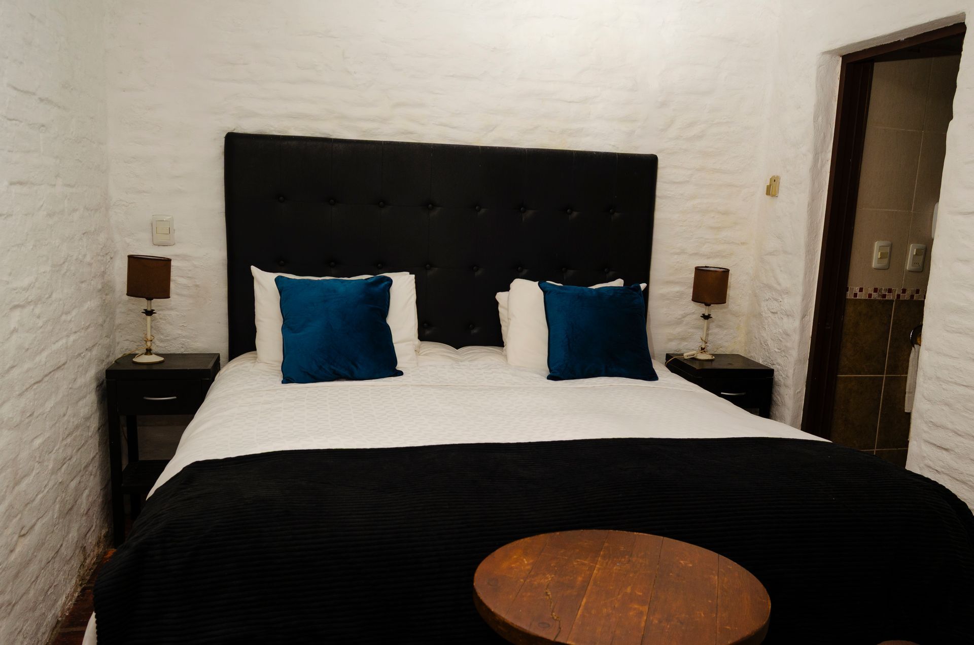 Un dormitorio con paredes de ladrillo blanco, un cabecero capitoné negro, una cama blanca con ropa de cama negra, dos almohadas azules y lámparas.