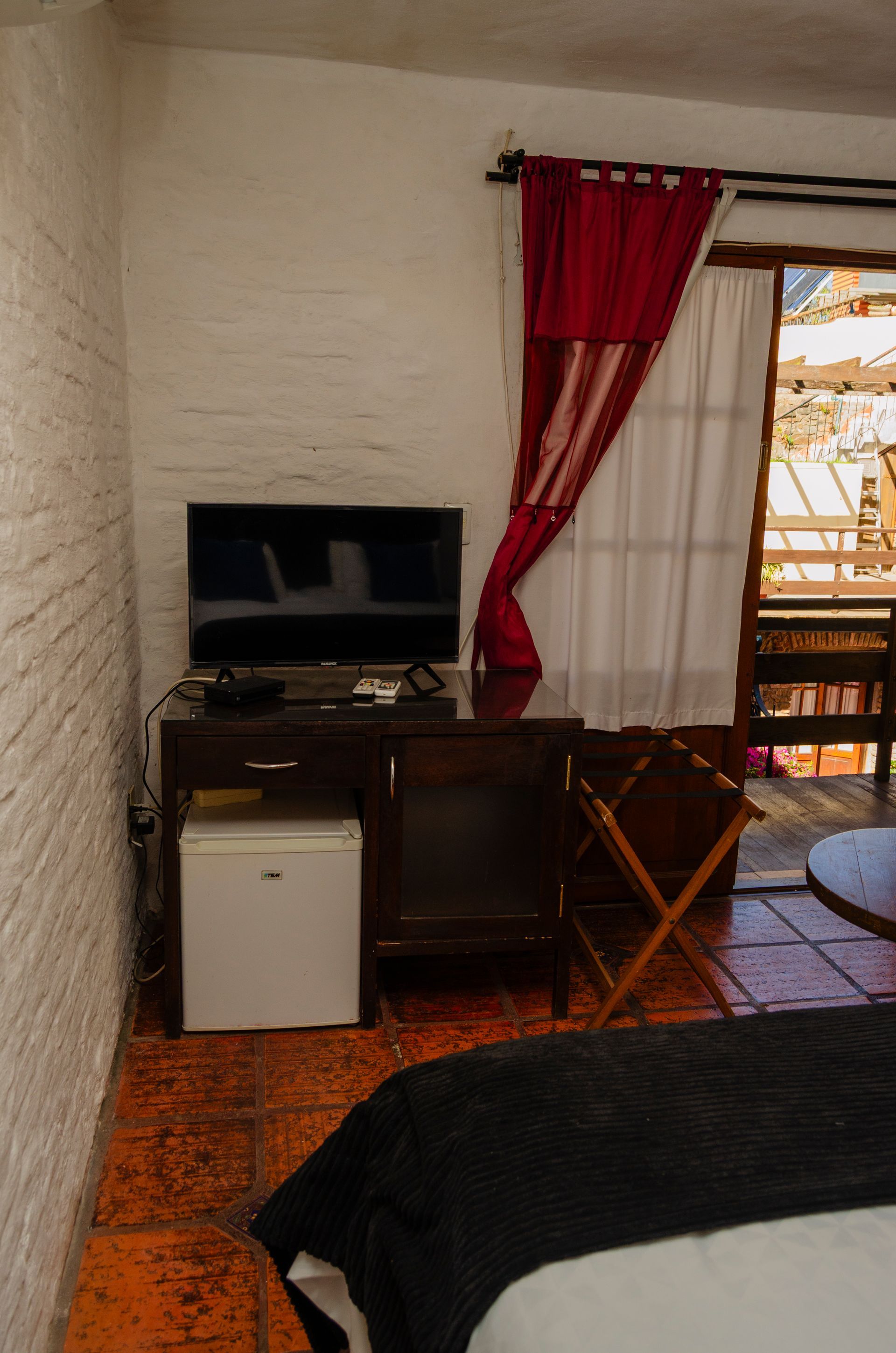 El interior de un dormitorio cuenta con un televisor sobre un soporte de madera, un mini refrigerador blanco, cortinas rojas y suelo de baldosas de terracota.