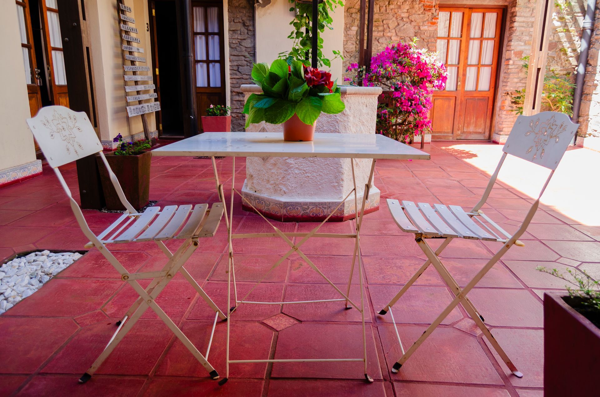 Un conjunto de mesa y sillas de bistró con dos sillas de metal blanco y una mesa cuadrada, situado en un patio con baldosas rojas, plantas y elementos arquitectónicos de piedra.