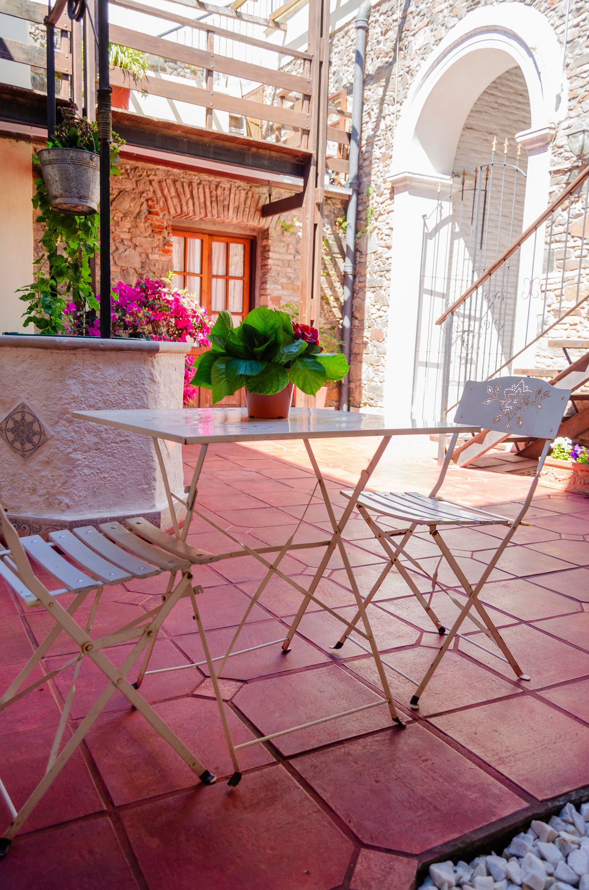 Una mesa de bistró y dos sillas plegables en un patio con baldosas rojas, con una estructura de piedra, plantas en macetas y un arco detrás.