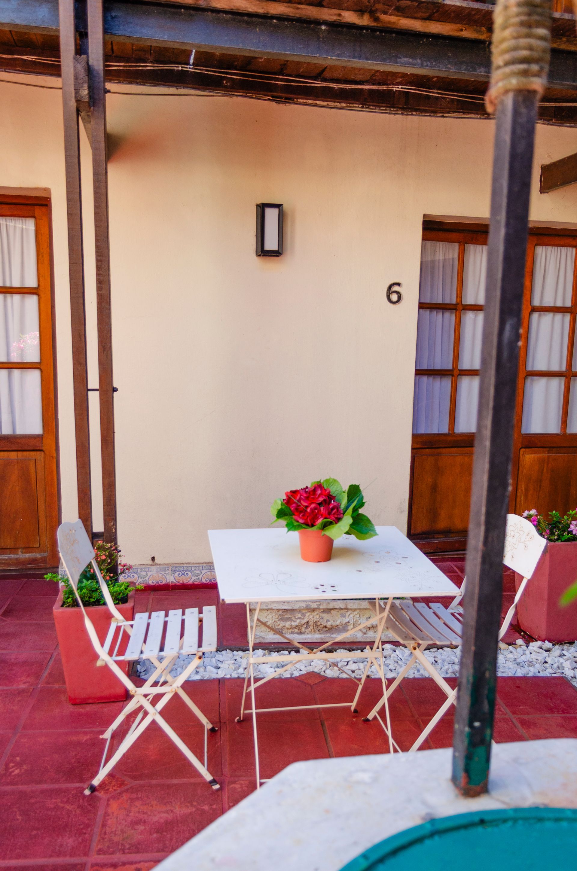 Un conjunto de mesa y sillas de bistró blancas con una pequeña maceta roja se encuentra en un patio con baldosas rojas frente a un edificio con la habitación número 6.
