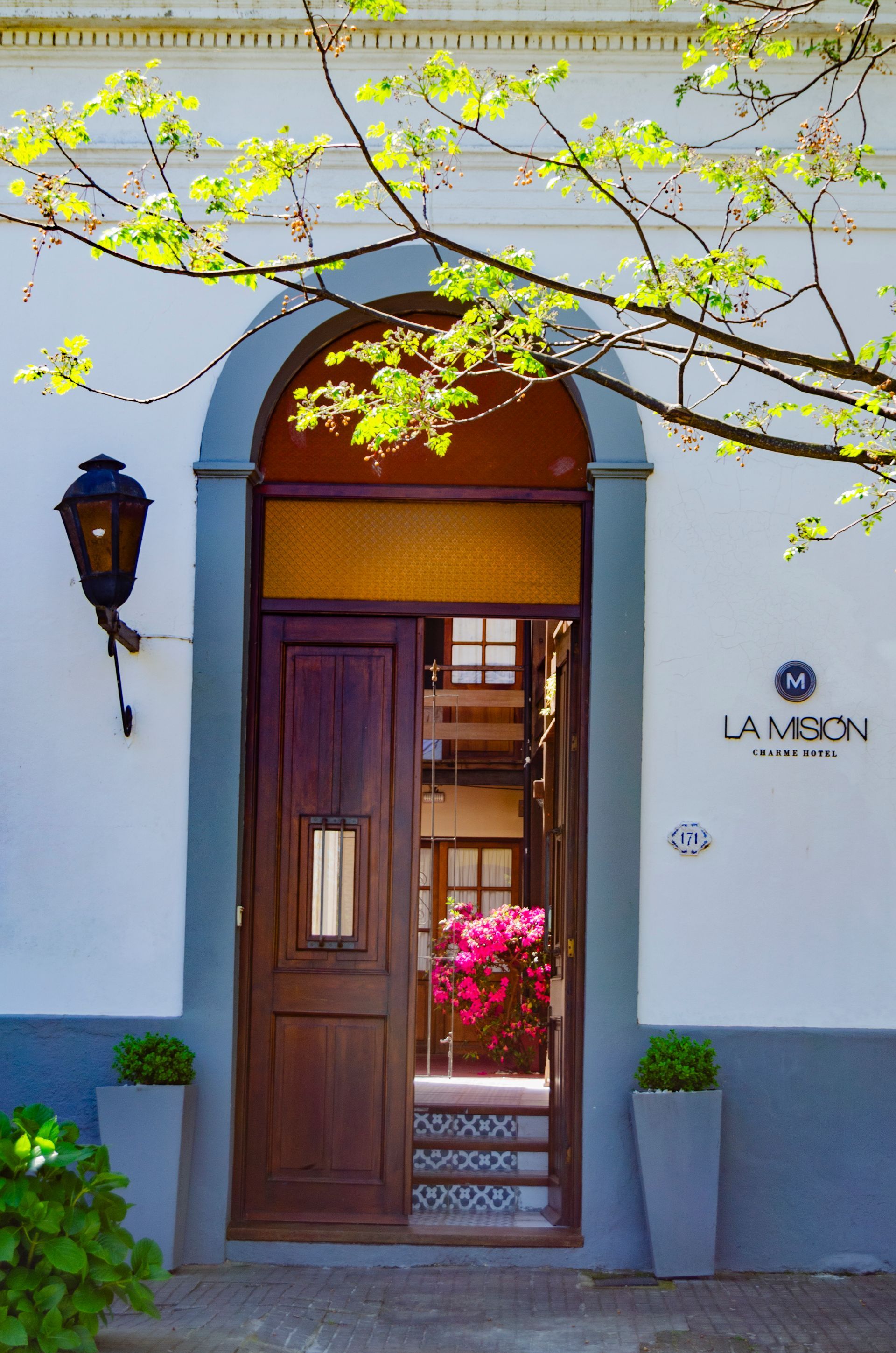 Entrada de madera arqueada en La Mibon, flanqueada por jardineras grises y una linterna de pared, bajo las ramas de un árbol frondoso.