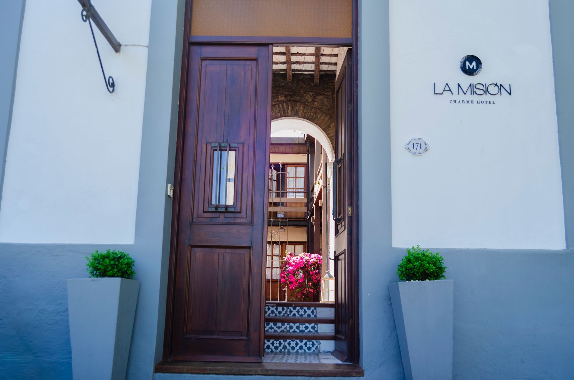 En el Hotel La Misión, una puerta de madera da acceso a un patio al aire libre con escaleras decoradas y flores rosas.