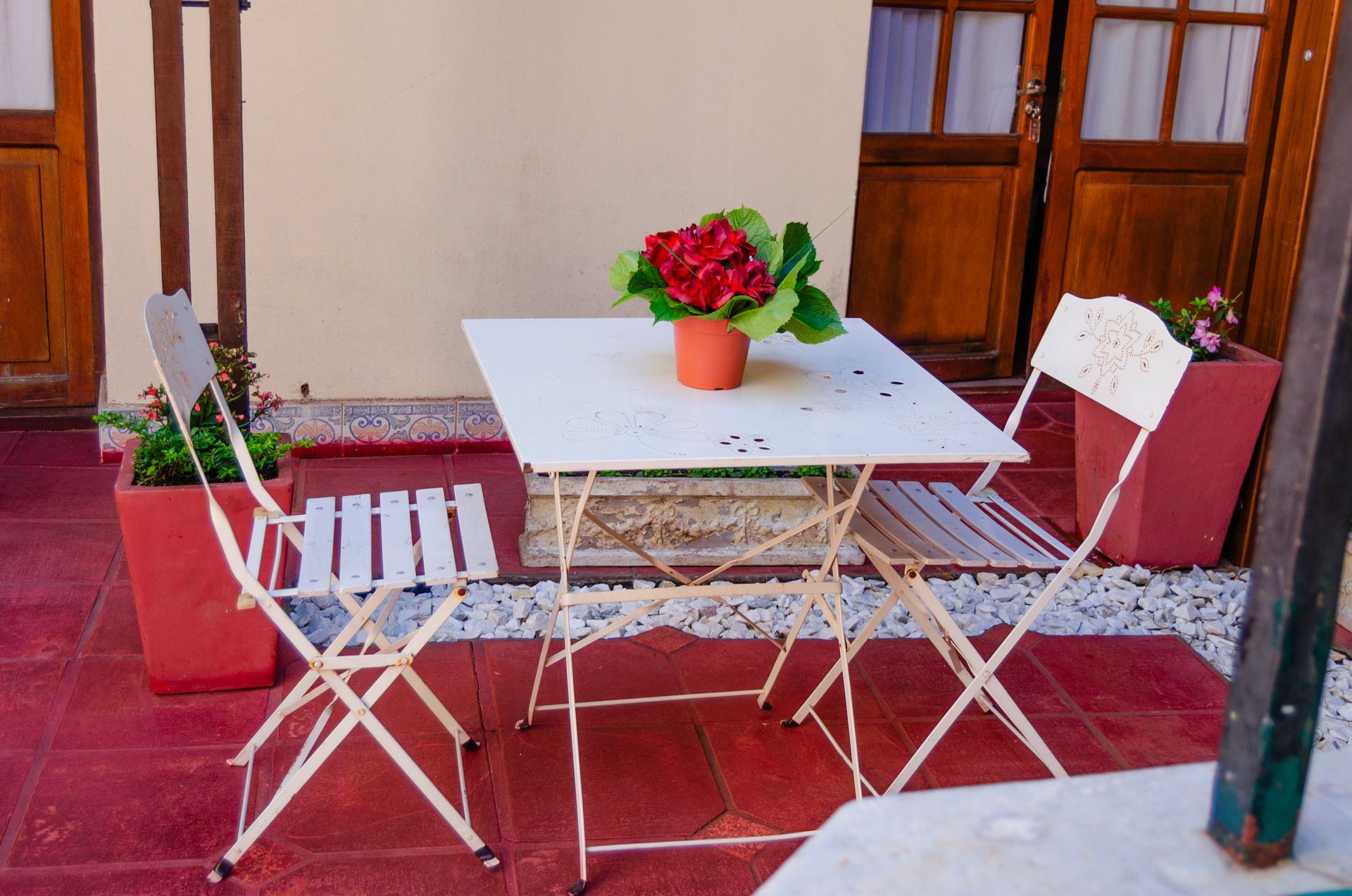 Un conjunto de mesa y sillas de bistró blancas, con una mesa cuadrada coronada por una maceta con una flor roja, se encuentra en un patio con baldosas rojas.