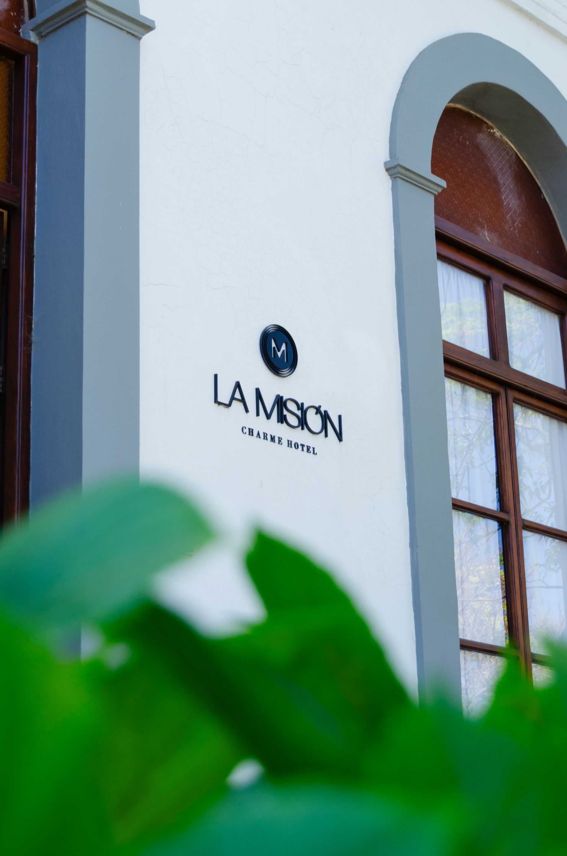 El logotipo del Hotel Boutique La Misión sobre una pared blanca, enmarcado por hojas verdes difuminadas y una ventana arqueada.