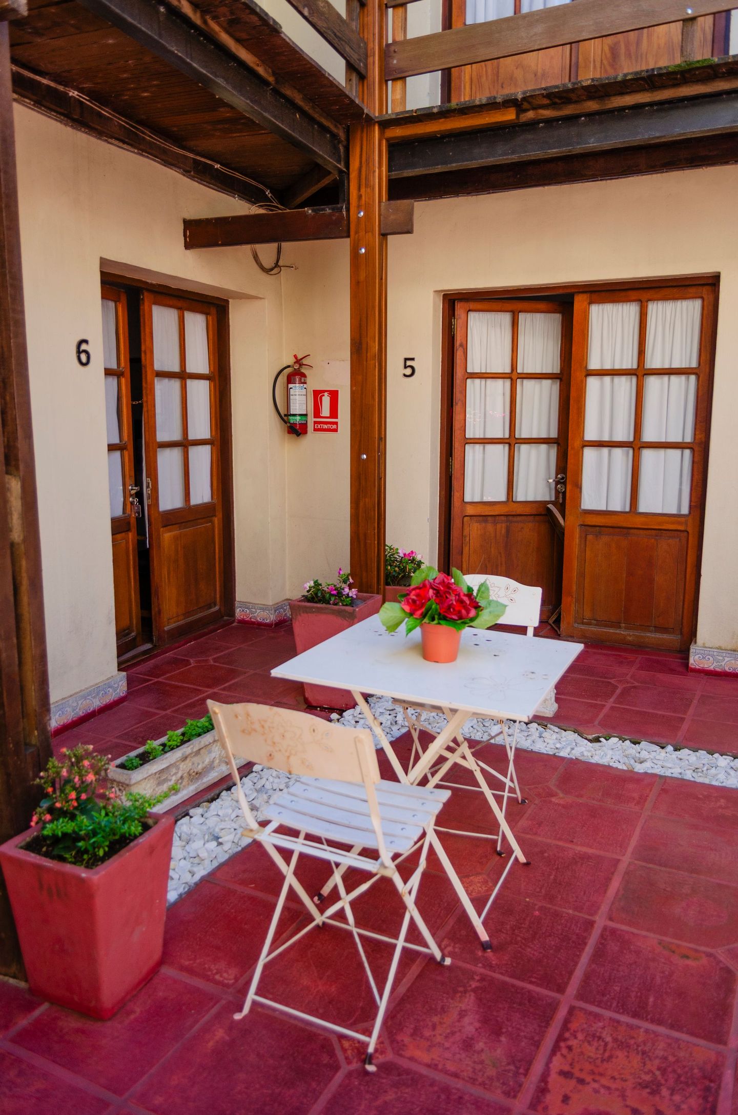 Un patio con suelo de baldosas rojas, una mesa de metal blanca con flores y dos puertas con los números 5 y 6.