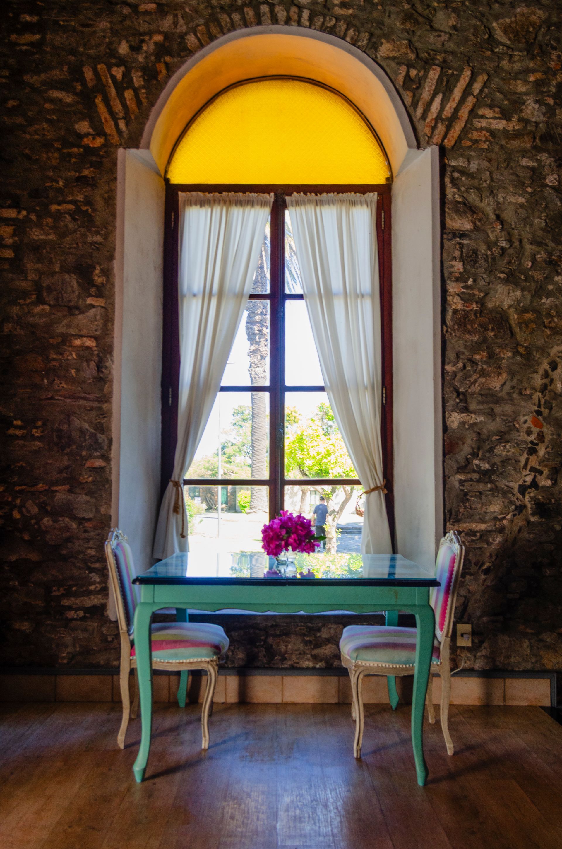 Una mesa de comedor con dos sillas se sitúa frente a un gran ventanal arqueado, adosado a una pared de piedra, y enmarcado por unas cortinas blancas translúcidas.