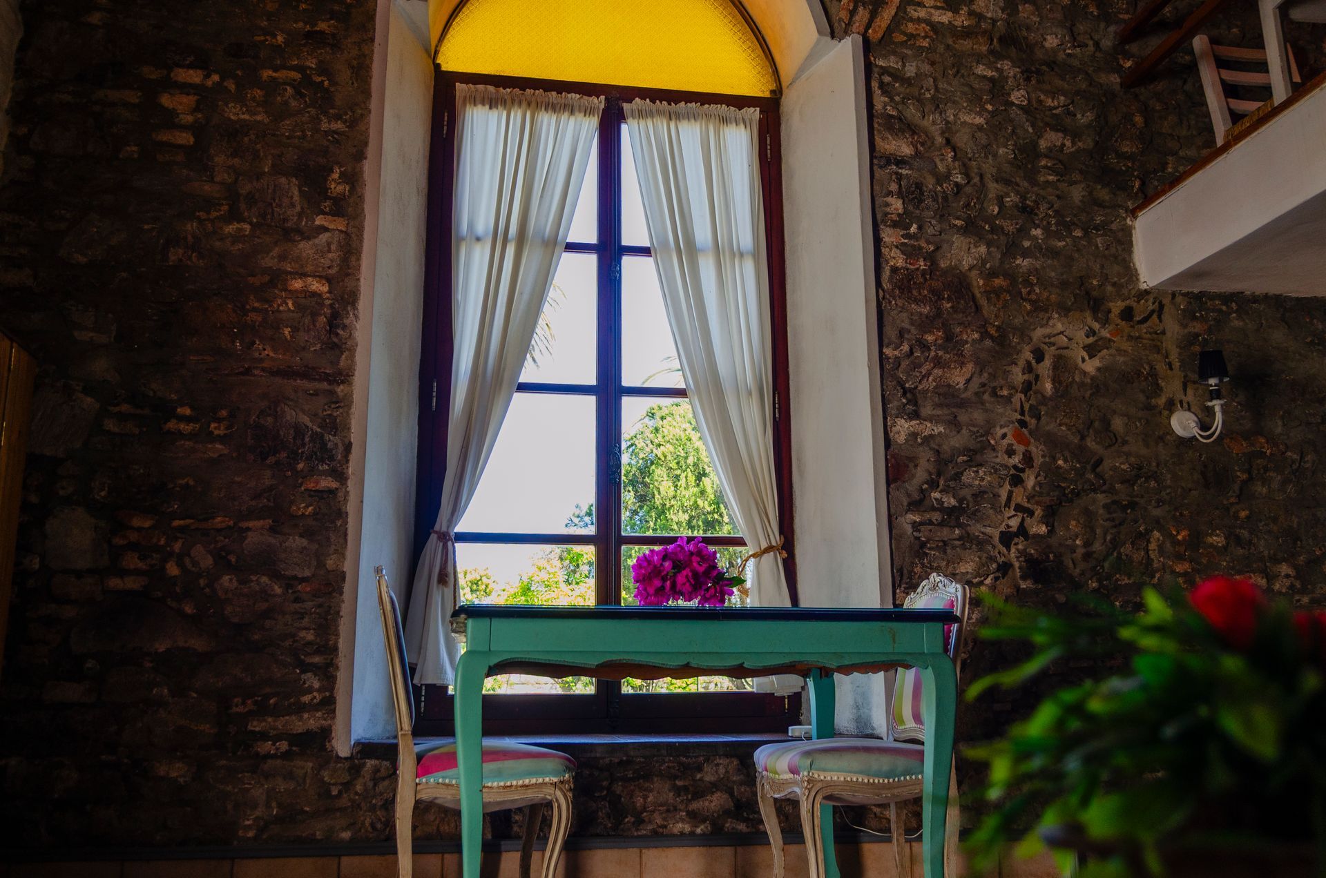 Una mesa de color turquesa y dos sillas se ubican frente a una gran ventana con cortinas blancas en una habitación rústica con paredes de piedra.
