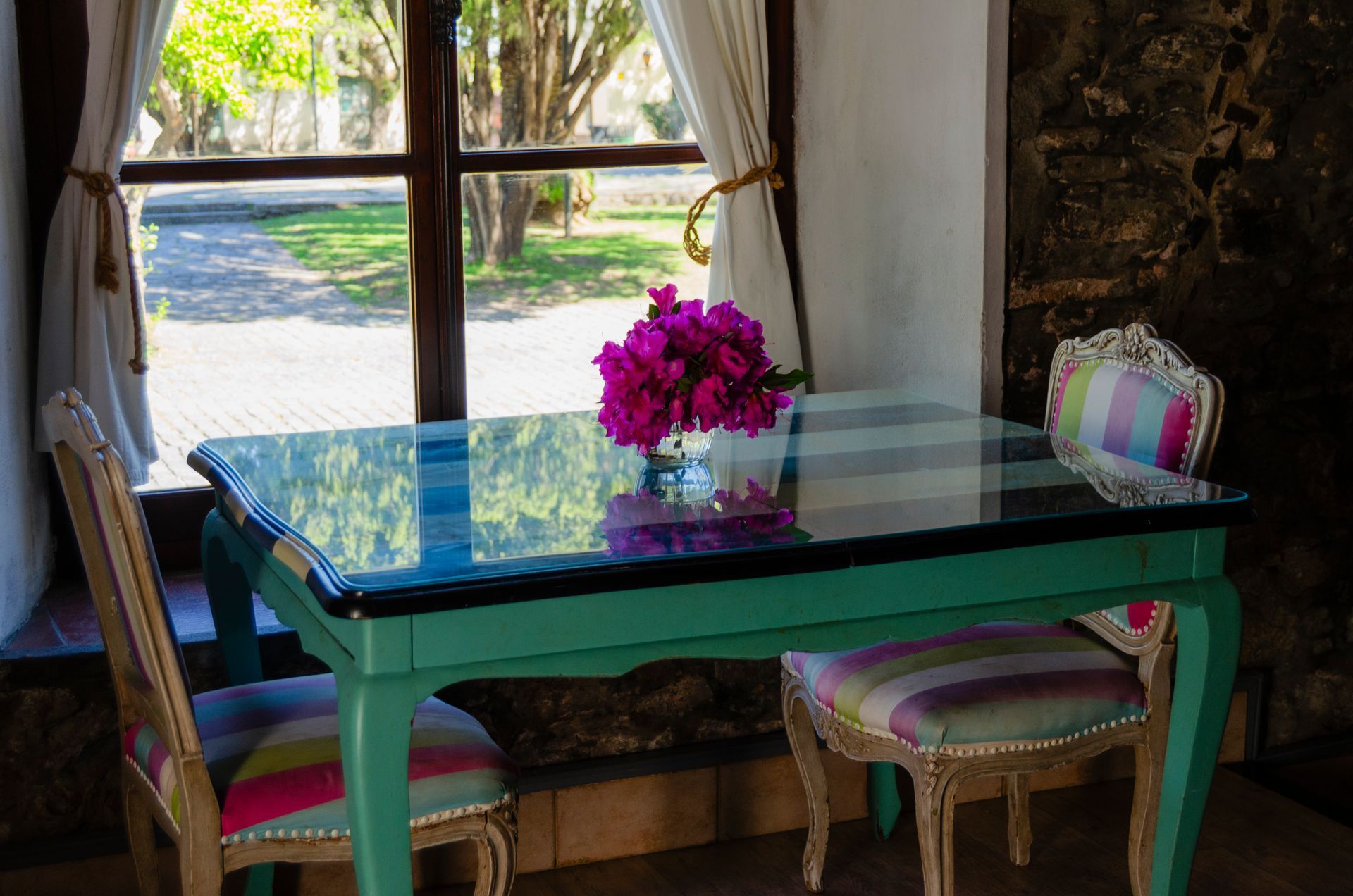 Una mesa de color verde azulado con un ramo de flores magenta se encuentra entre dos sillas con cojines de rayas multicolores, cerca de una ventana.
