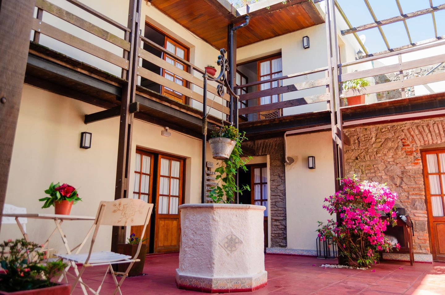 Un patio con un pozo de piedra, balcones de madera, una mesita con una planta en maceta y buganvillas de un vibrante color rosa.