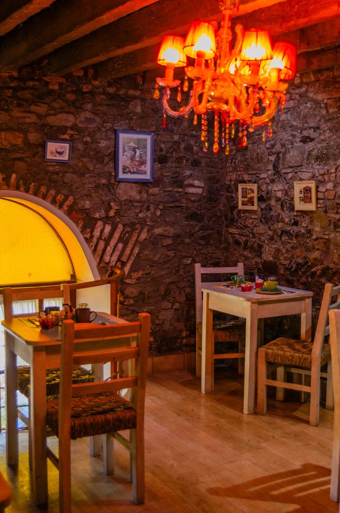 Una habitación rústica y acogedora con paredes de piedra, dos pequeñas mesas de comedor, sillas de madera y una lámpara de araña vibrante de tonos cálidos.