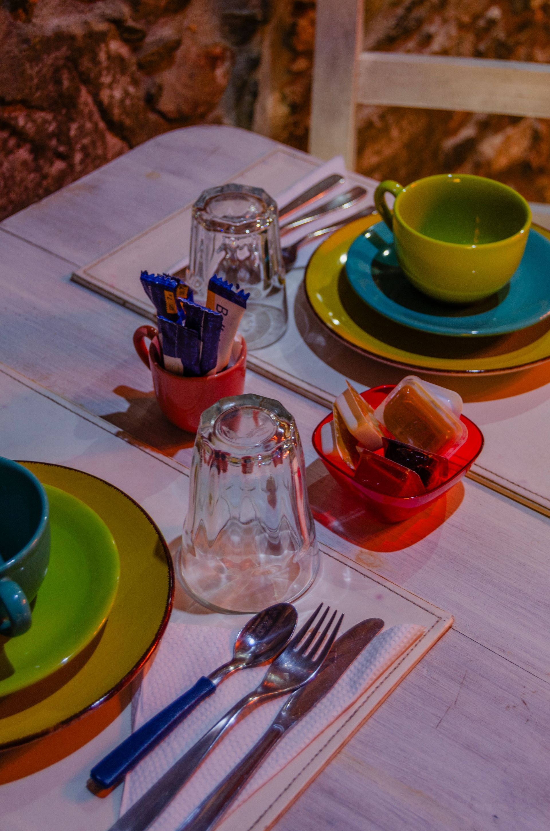 Una mesa puesta sobre madera con platos, tazas, vasos invertidos, cubiertos y pequeños cuencos con aperitivos de colores.
