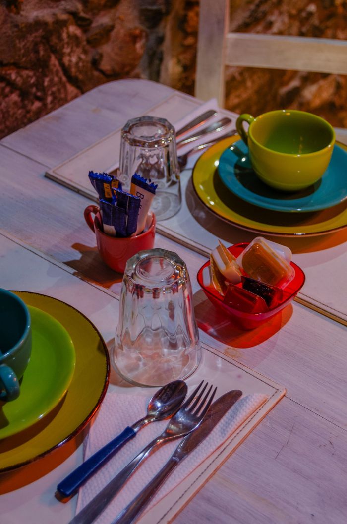 Una mesa puesta con dos juegos de cubiertos coloridos, vasos, cubiertos, un azucarero y un pequeño cuenco con aperitivos.