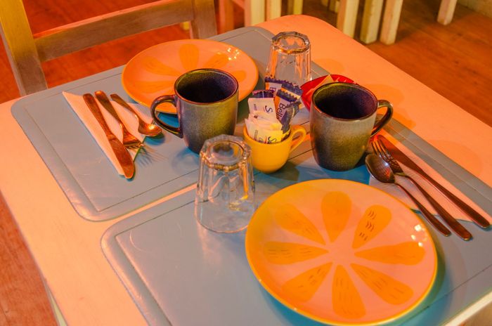 Una mesa puesta con dos platos con motivos cítricos de color naranja, tazas, vasos, cubiertos y sobres de azúcar.