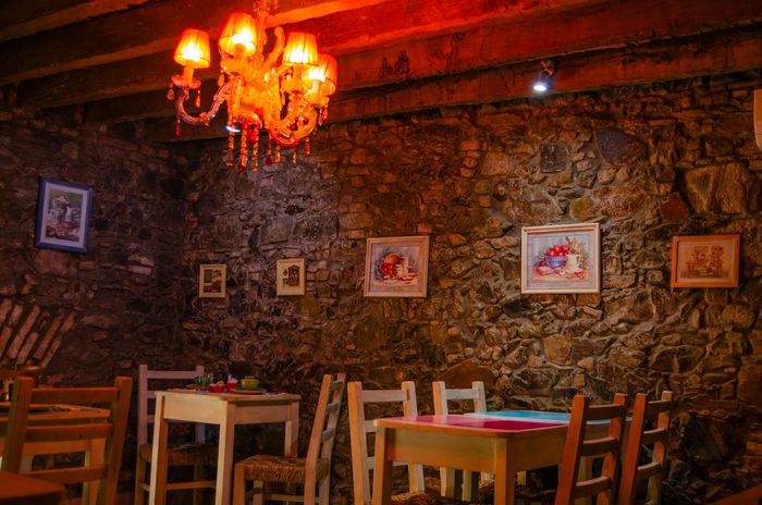 Comedor con iluminación tenue, paredes de piedra rústica, mesas y sillas de madera, y una lámpara de araña roja decorativa.