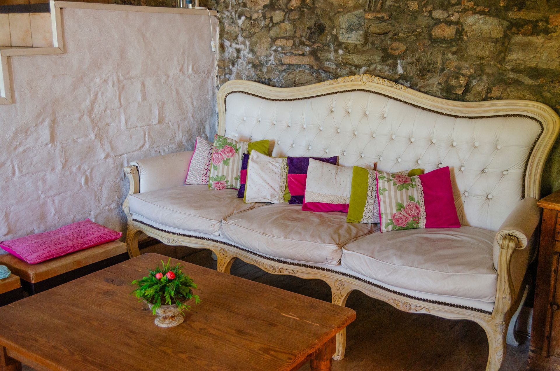 Un sofá blanco capitoné de estilo vintage, con cojines decorativos de colores, se encuentra en una habitación con paredes de piedra y una mesa de centro de madera.