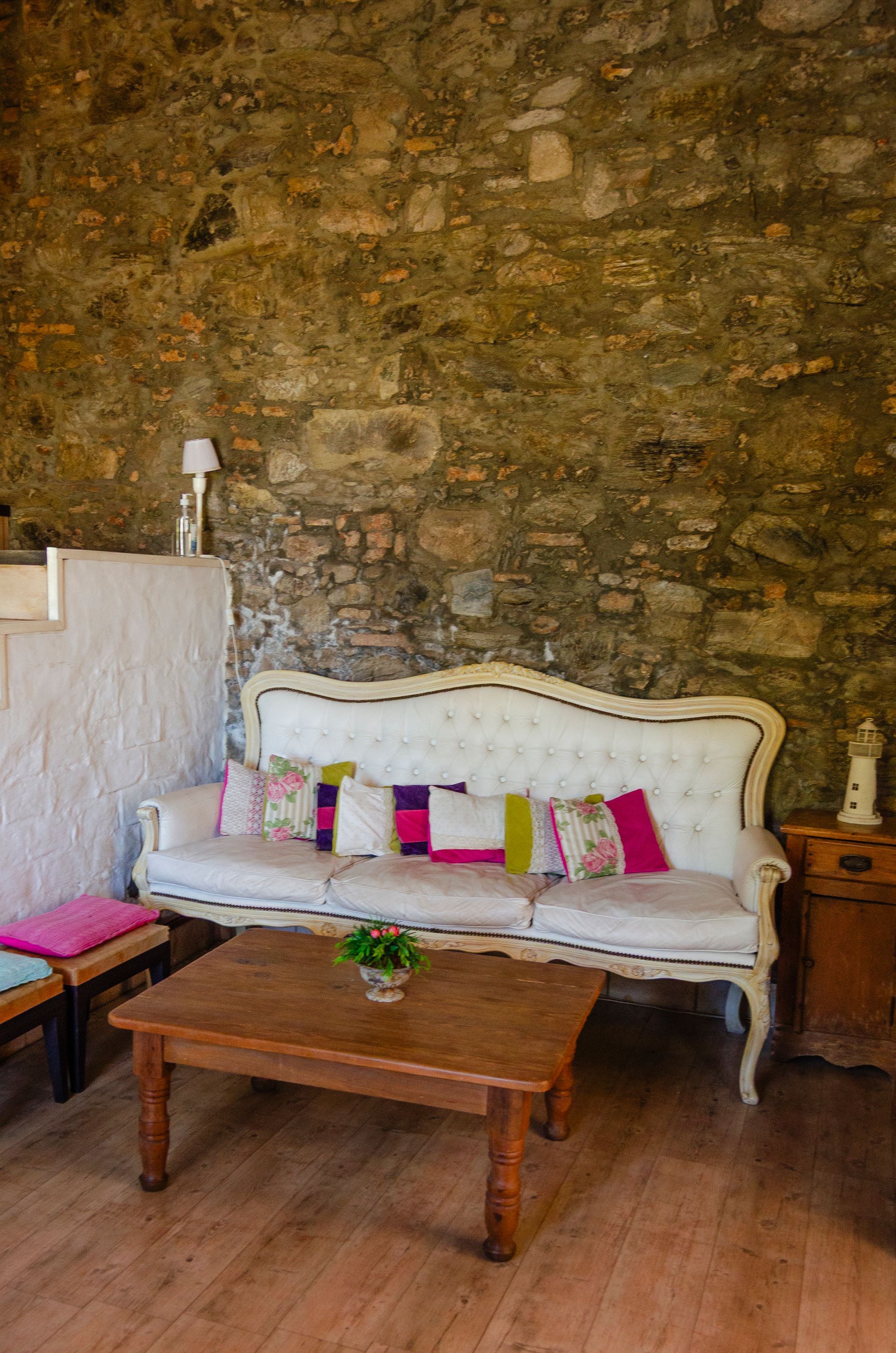 Un sofá blanco capitoné con cojines decorativos de colores se sitúa frente a una mesa de centro de madera, junto a una pared de piedra rústica.